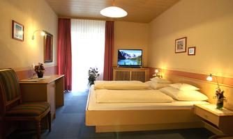 A cozy hotel room with two single beds and a television. The room features a bright wooden ceiling and large windows with a view outside.