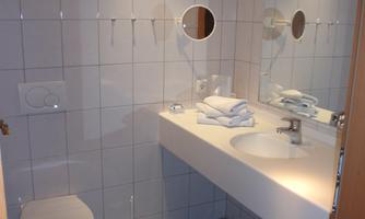 A modern bathroom with white tiles and a large mirror. There is a toilet, a sink, and some towels on the shelf.