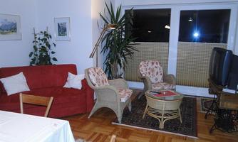 A cozy living room with a red sofa, a round table, and two upholstered chairs.  
Large windows let in plenty of light, and plants create an inviting atmosphere.