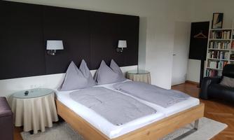 A modern bedroom with a large double bed and elegant bedside lamps. The walls are designed more simply, and an armchair and a bookshelf are visible in the background.