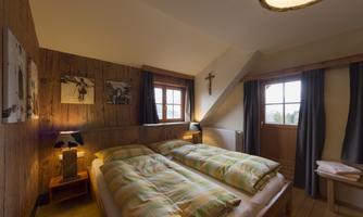 A cozy bedroom with two beds, wooden paneling, and pictures on the wall. Windows let in plenty of light and create an inviting atmosphere.