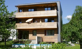 A modern house with a wooden facade and several balconies. The surroundings consist of green grass and trees under a clear blue sky.