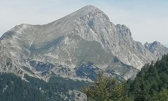 A majestic mountain with steep rocks and green forests in the foreground. The sky is clear and blue.