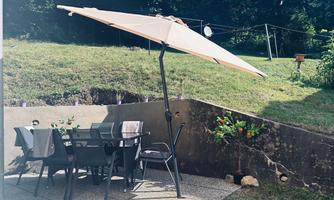 A cozy outdoor area with a large umbrella and a table with chairs. In the background, the garden with a small plant is visible.