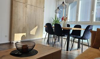 A modern living room with a wooden table and black chairs. Large windows let in plenty of light, and plants create a fresh atmosphere.