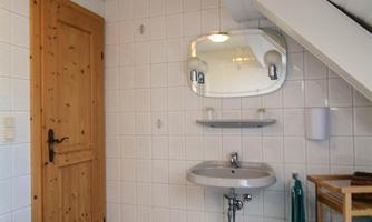 A small, modern bathroom with white tiles and a wooden shelf. The wall is decorated with a large mirror and a lamp.
