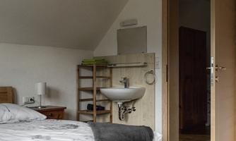 A simple bedroom with a wooden bed and a cozy blanket. In the background, there is a sink and an open door to another room.