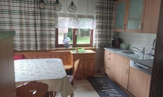 A cozy kitchen with wooden cabinets and a large window. A white tablecloth is on the table, and the floor is decorated with a rug.
