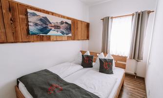 A cozy bedroom with a double bed and wood paneling. The windows are adorned with curtains, and above the bed hangs a landscape painting.