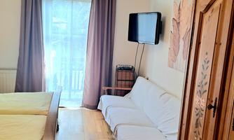 A cozy bedroom with two beds, a sofa, and a TV. Large window with curtains and warm wooden floor.