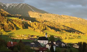 A picturesque landscape with green hills and a small church in the foreground. The sun illuminates the mountains in the background.