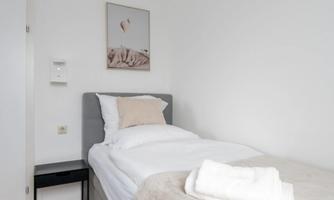 A simple bedroom with a single bed and a gray bedspread. A picture hangs on the wall, and there is a small nightstand.