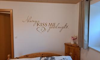 A bedroom with a wooden bed and white bedding. On the wall, there is a decorative inscription: "Always KISS ME goodnight."