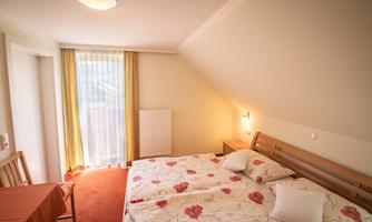 A bright bedroom with a double bed and colorful floral patterns on the bedding. There is a small table and a balcony with sheer curtains.