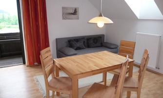 A cozy living area with a wooden table and four chairs. A gray sofa and large windows provide plenty of light.