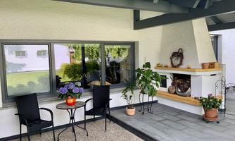 A cozy outdoor area with two chairs and a small table. In the background, there are plants and a fireplace.