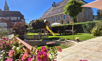 A beautiful garden with colorful flowers, a playground slide, and an inviting terrace. In the background, there is a large house under a blue sky.