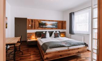A modern bedroom with a double bed and wooden details. There is a table and large windows that let in plenty of light.