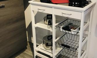 A white kitchen cart with kitchen appliances like a coffee maker and a kettle. Below are some pots on shelves.
