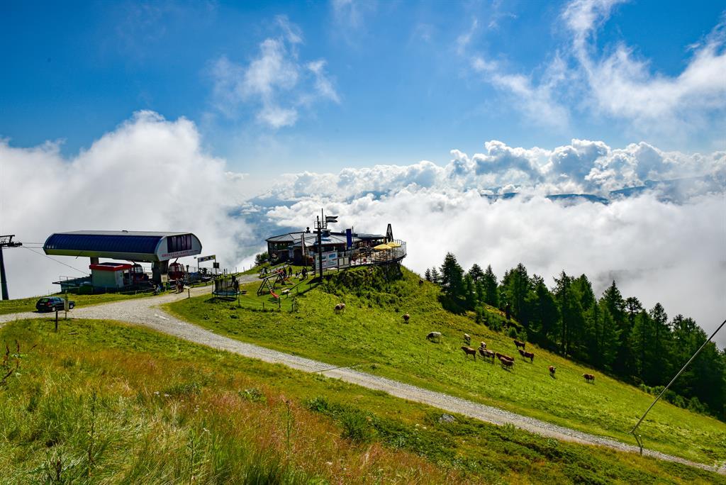 A picturesque mountain landscape with a cable car station and a meadow full of cows. The sky is clear with some clouds hanging over the mountains.
