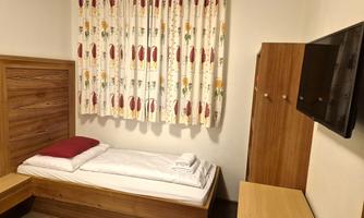 A simple room with a single bed, a table, and a television. The curtain has a colorful pattern and there are fresh towels on the bed.
