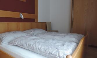 A simple bedroom with a wooden bed and scattered pillows. The room has a calm atmosphere with a wardrobe and a chair.