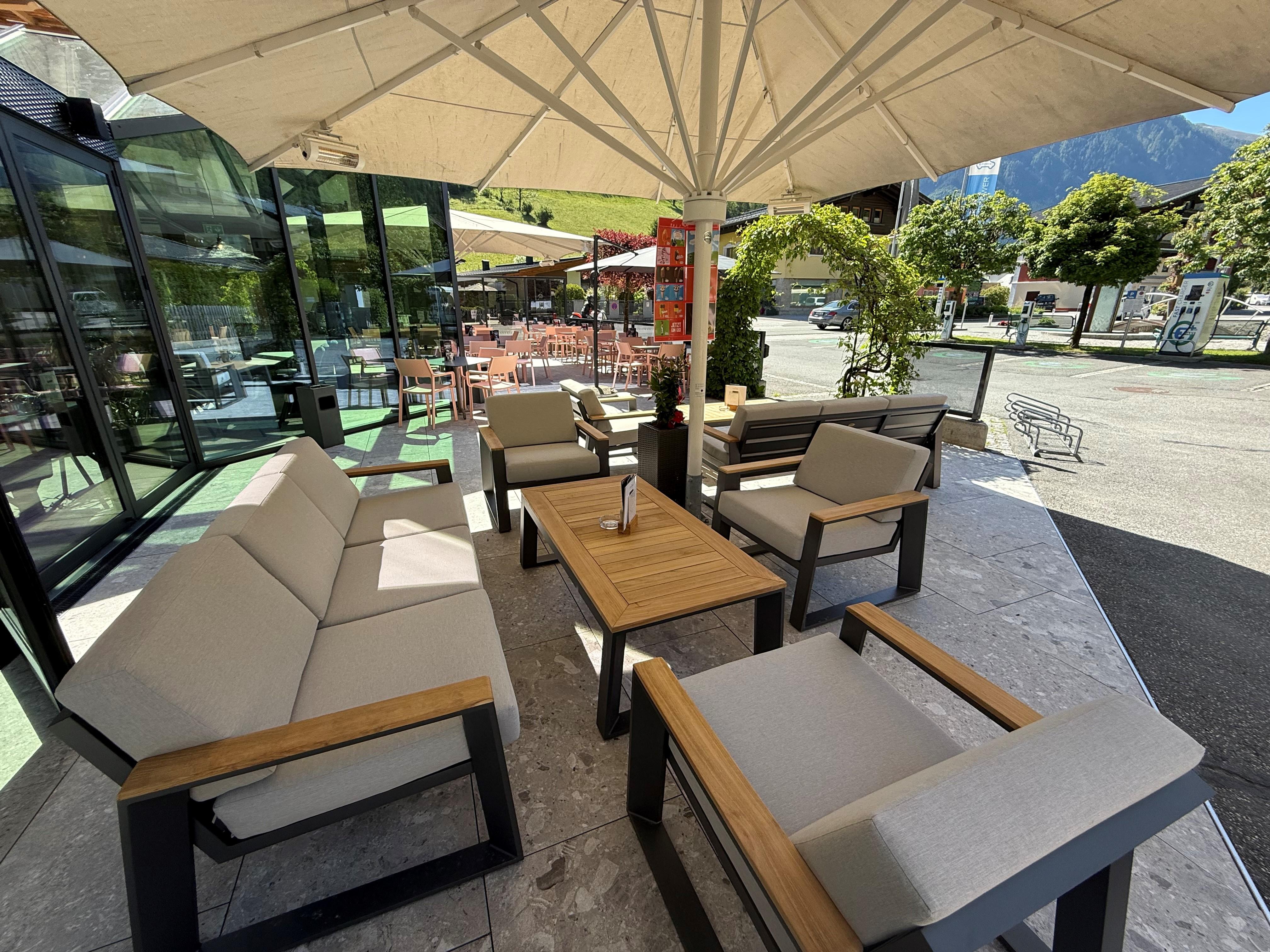 A cozy seating area under a sunshade with modern furniture. In the background, there is an outdoor area with a view of nature.