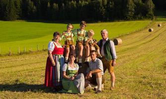 A family in traditional costumes stands in a green field. In the background, trees and hay bales can be seen.