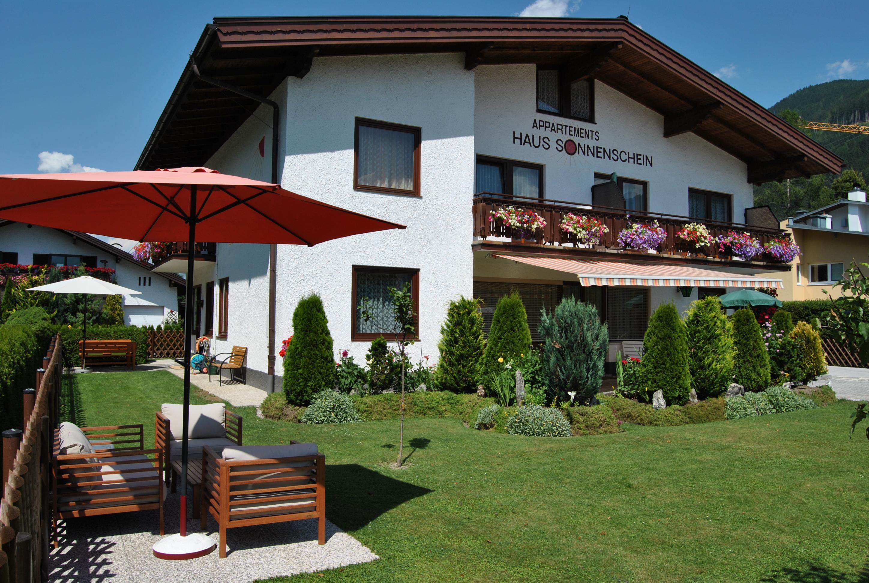 A charming house with a beautiful garden and a cozy sitting area. Red umbrellas and blooming plants create an inviting atmosphere.