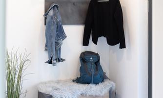 An inviting hallway with a wall shelf on which clothes hang. Below is a bench with a cushion and shoes beside it.