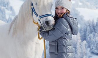 A girl is standing next to a white horse in the snow. In the background, snowy mountains and trees can be seen.