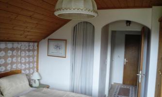 A bright bedroom with a wooden ceiling and a large window. The room is cozily furnished with a bed and a lamp.