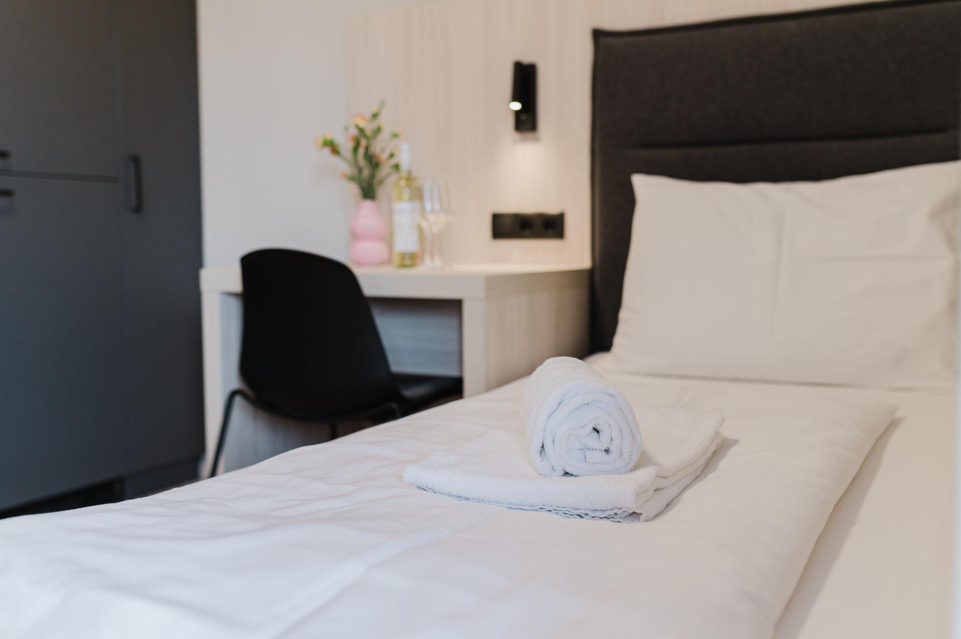 A cozy room with a single bed, fresh towels, and a small table. On the table, there are flowers and a bottle of water.