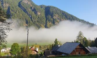 A tranquil mountain landscape with fog rolling over the valley. In the foreground, traditional wooden houses and green meadows are visible.