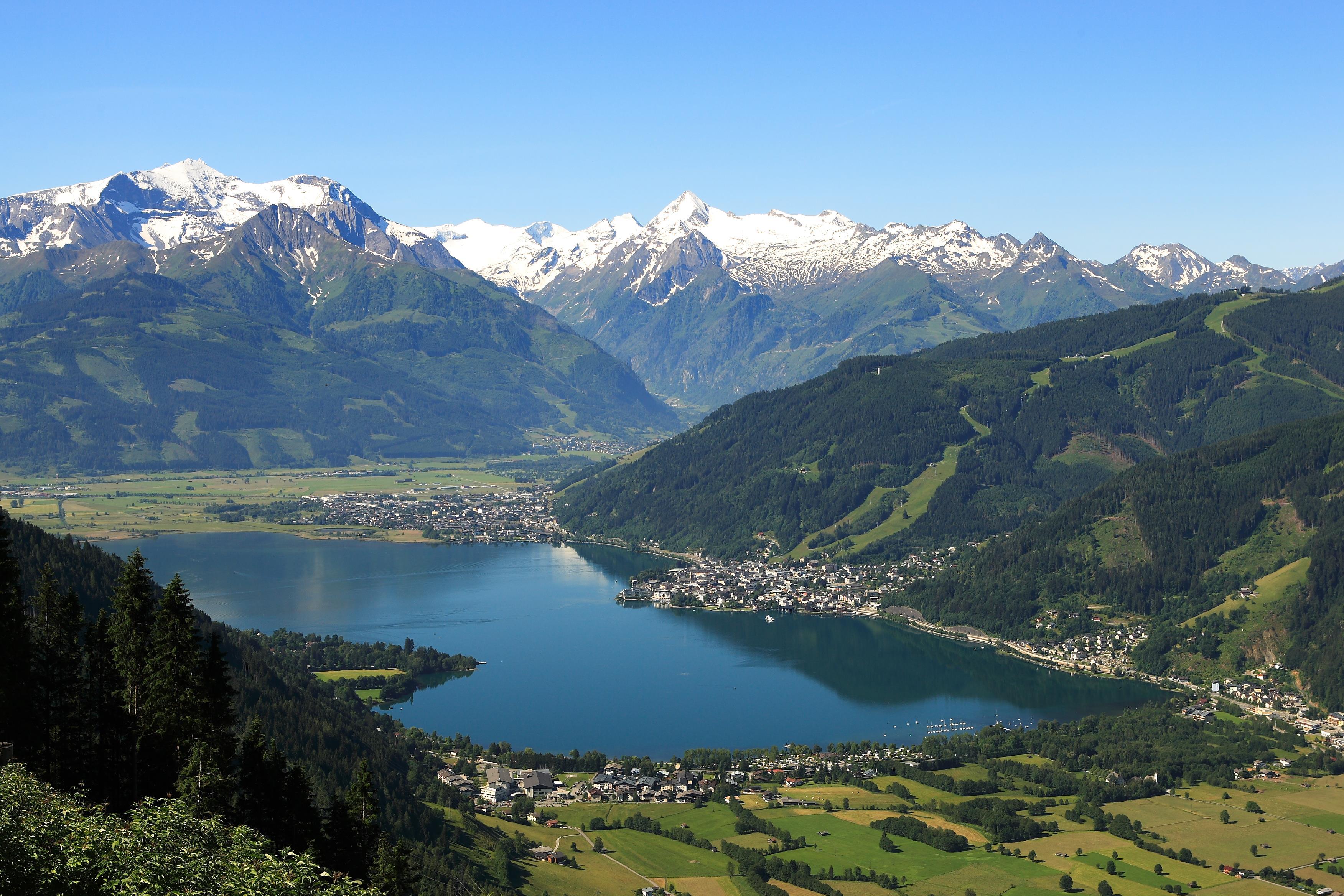 An impressive mountain landscape with snow-capped peaks and a clear blue lake. In the foreground, green meadows and a picturesque village can be seen.