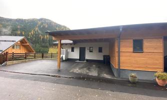 A modern house with a wooden facade and a covered terrace. The parking lot is paved and surrounded by green mountains.