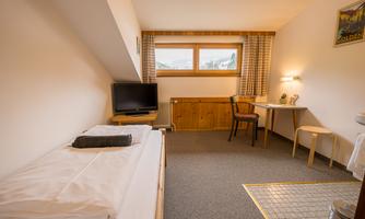A simple room with a bed, a television, and a small table with a chair. The room has a window with an outside view and a warm atmosphere.