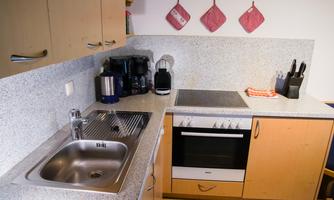 A modern kitchen with a sink, stove, and various kitchen appliances. The walls are decorated with gray stone and red pot holders.