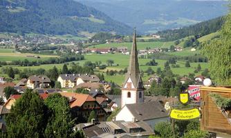A picturesque landscape with green hills and a charming village. In the foreground, a church tower is visible, surrounded by traditional buildings.