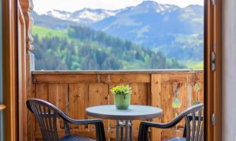 A balcony with two chairs and a small table. In the background, mountains and a green landscape can be seen.