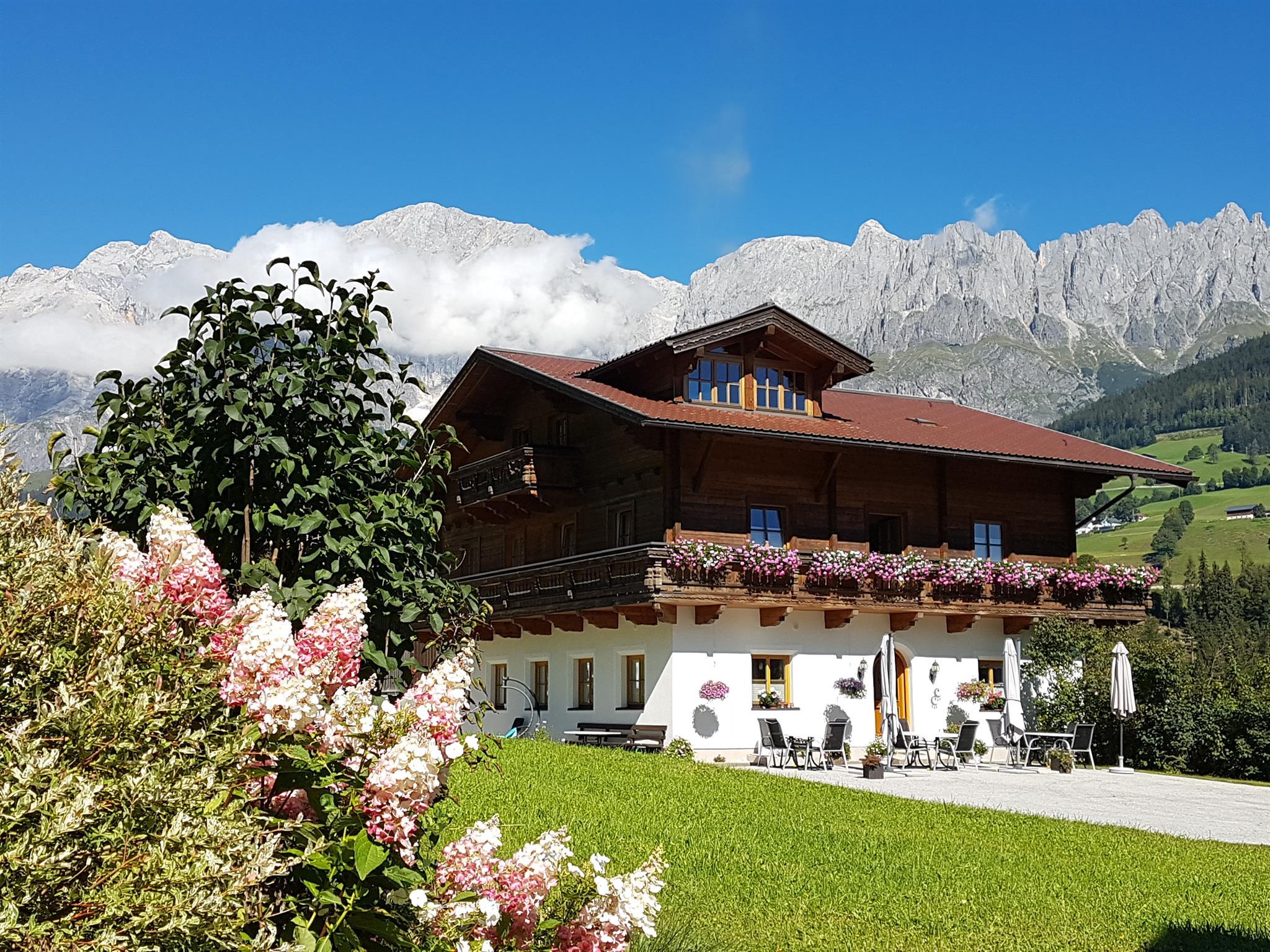 A picturesque alpine house with flowers on the balcony. In the background, majestic mountains rise under a clear blue sky.