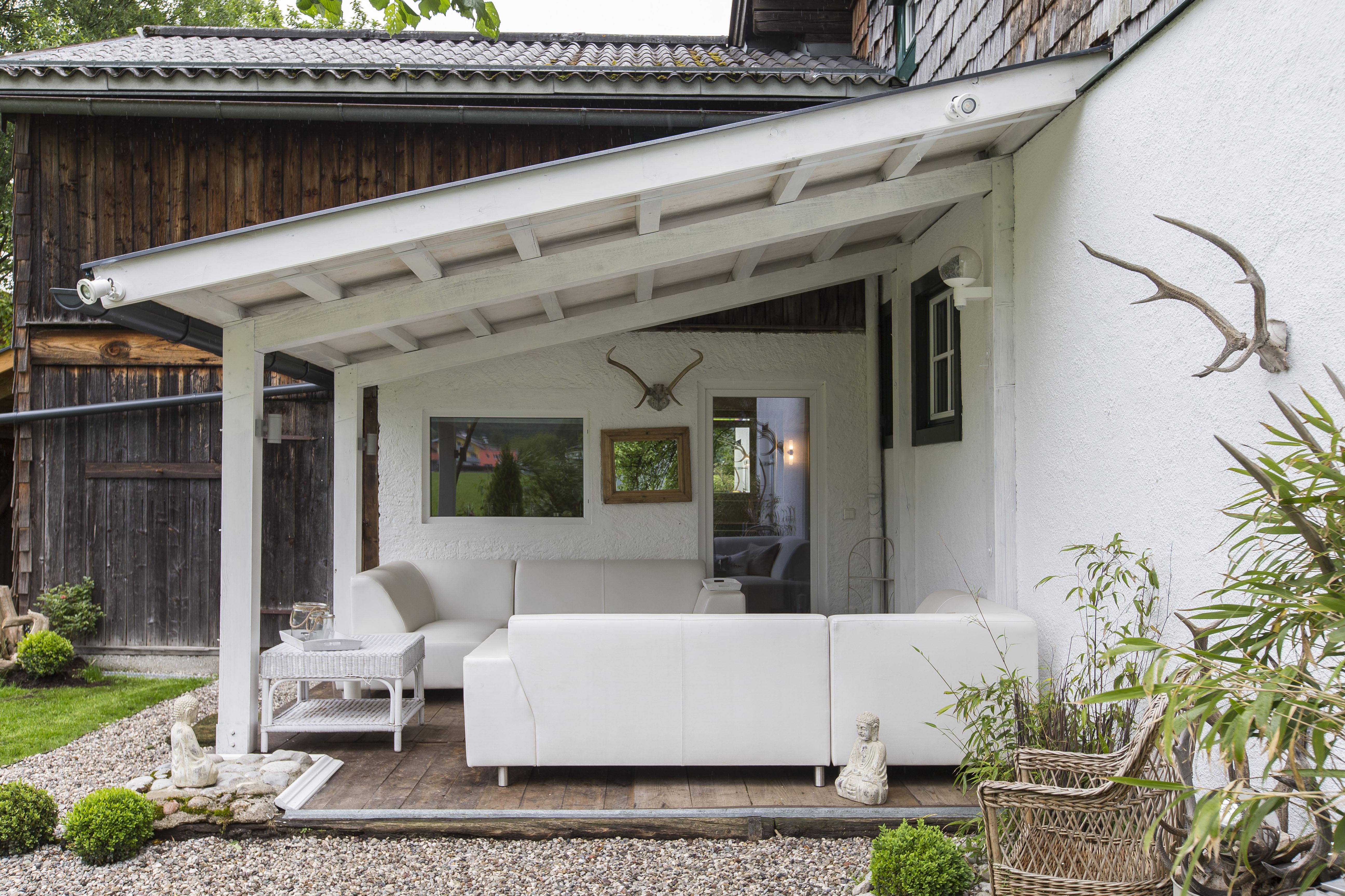 A cozy terrace with a white sofa and a covering. The area is surrounded by green plants and gravel.