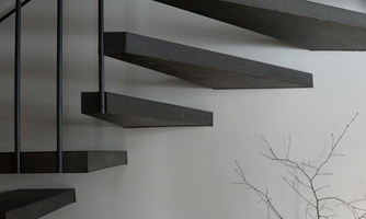 A modern spiral staircase made of wood with delicate metal railings. In the foreground stands a simple vase with a thin branch.