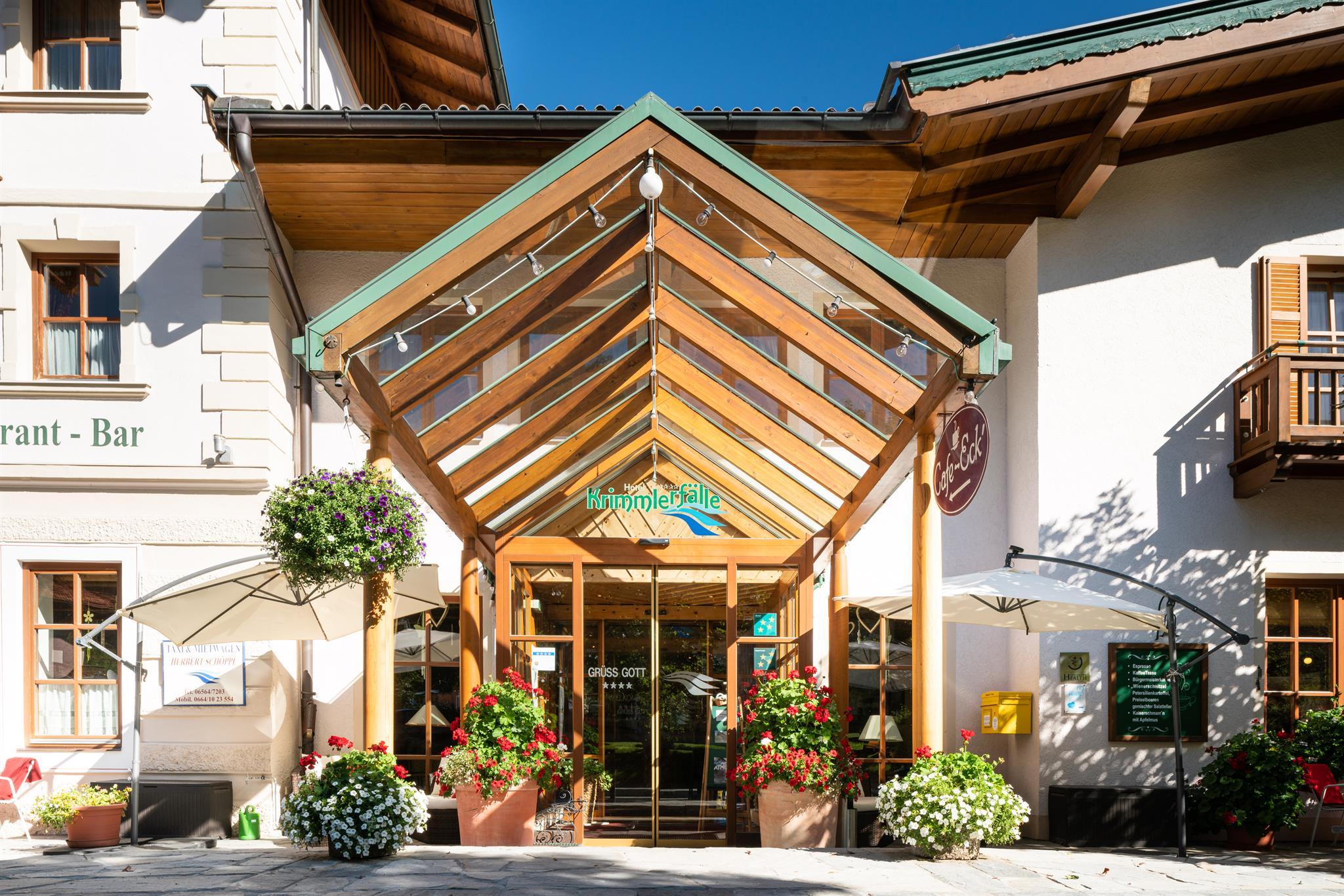 An inviting entrance area of a restaurant with wooden details and large windows. Surrounded by blooming plants and sun umbrellas, it offers a friendly atmosphere.