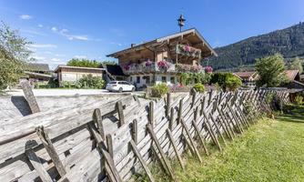 A traditional alpine house with colorful flowers and a wooden fence. In the background, green mountains and a blue sky can be seen.