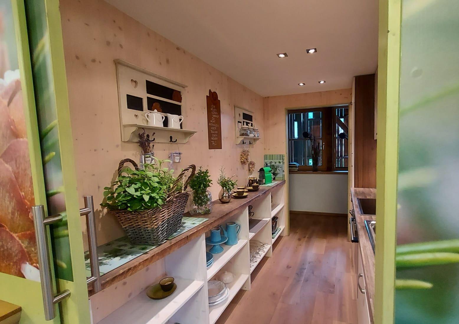 A cozy kitchen with bright walls and wooden flooring. Shelves with dishes and plants give the room an inviting atmosphere.