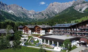 A picturesque hotel complex in the mountains with modern buildings and a green environment. Majestic mountains can be seen in the background and the sky is clear.