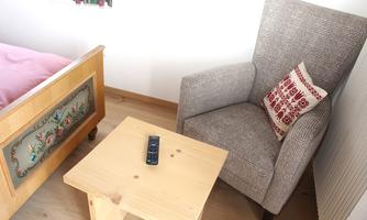 An armchair is next to a small wooden table. On the table lies a remote control, and in the background, there is a bed with decorative bedding.