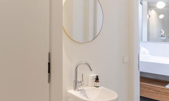 A modern bathroom with a white sink and a round mirror. In the background, a bright bathtub is visible.