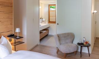 A modern and bright room with a cozy armchair and a small table. One can see a bathroom with a stylish sink and wooden details.